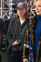 A woman wearing a dark striped button-up shirt, dark jeans, a gray baseball cap, and a brown shoulder bag stands outdoors next to another person in a patterned coat.