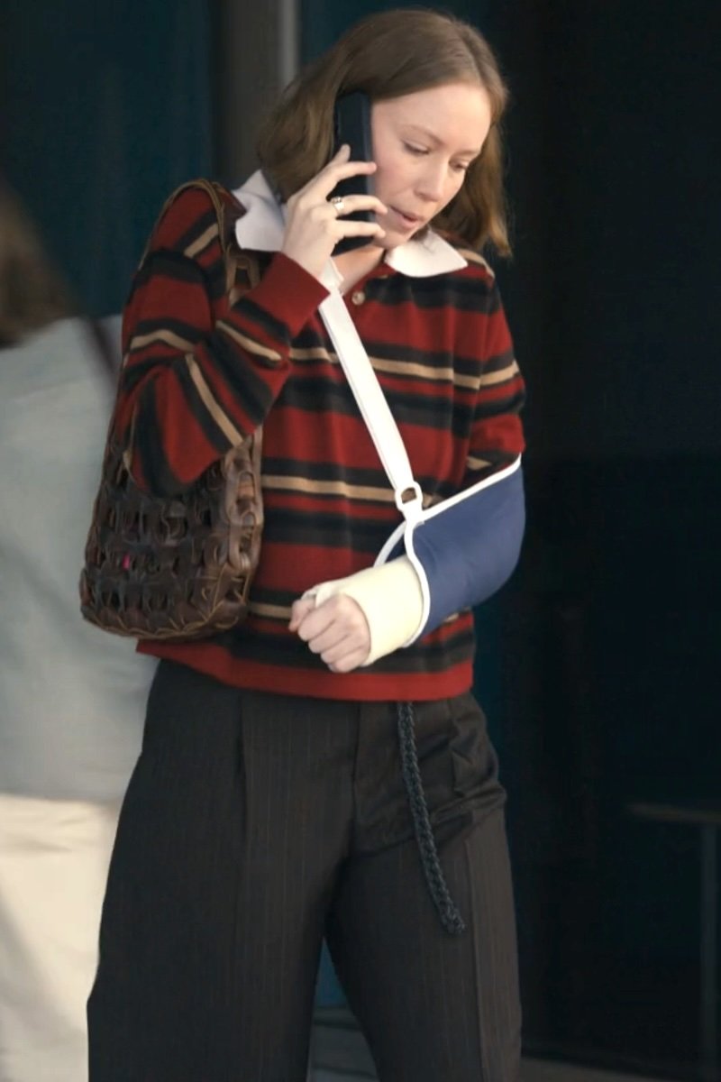 A woman wearing a red and black striped knit sweater with a white collar, dark pinstripe trousers, a brown woven shoulder bag, and a blue arm sling while talking on a phone.