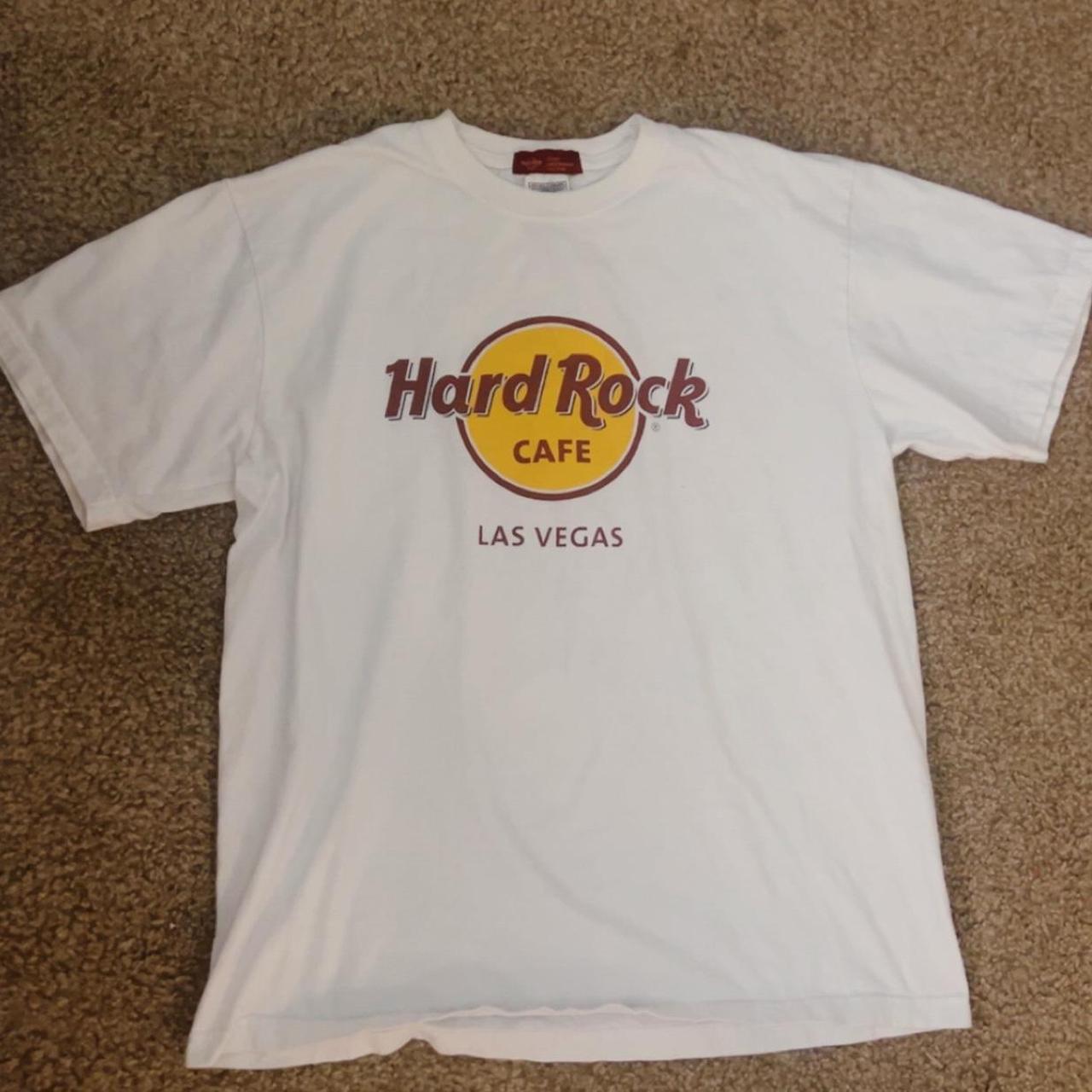 A white short-sleeve t-shirt with a Hard Rock Cafe Las Vegas logo printed on the front, laid flat on a brown carpeted floor.