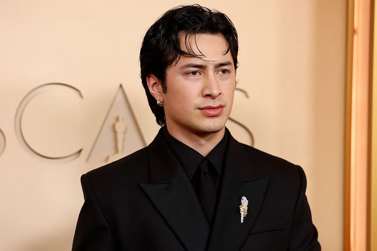 Man in a black suit with a brooch standing in front of an 'Oscars' backdrop