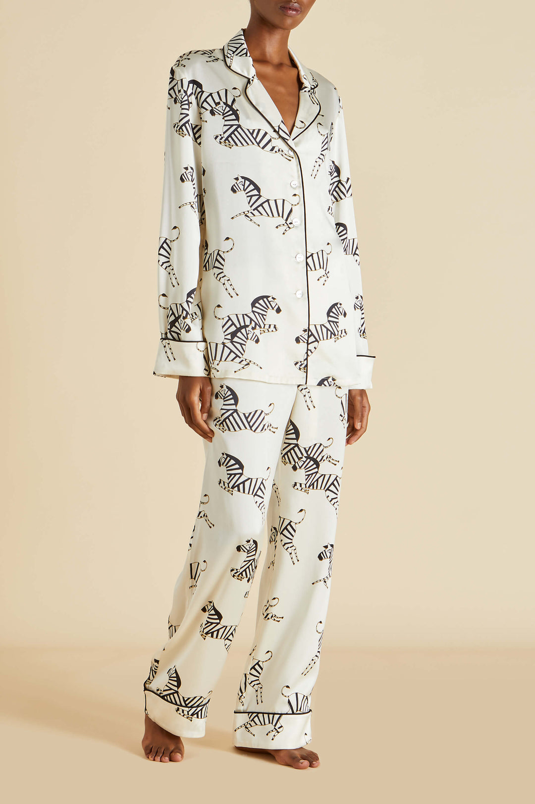 A model wearing a two-piece pajama set with a black and white zebra print on a cream background, standing against a solid beige backdrop.