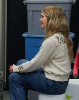Person wearing a cream-colored sweater with floral embroidery sitting on a white container.