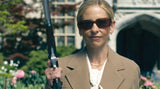 Woman holding a weapon in an outdoor setting with greenery and a building in the background