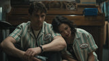 A man and a woman wearing matching green and white striped diner-style shirts with 'Bessie's' patches, seated indoors in a dimly lit restaurant or cafe setting.