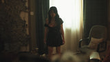 A woman in a short black dress stands in a dimly lit room near a bright window with curtains, with a chair and textured wall visible in the background.