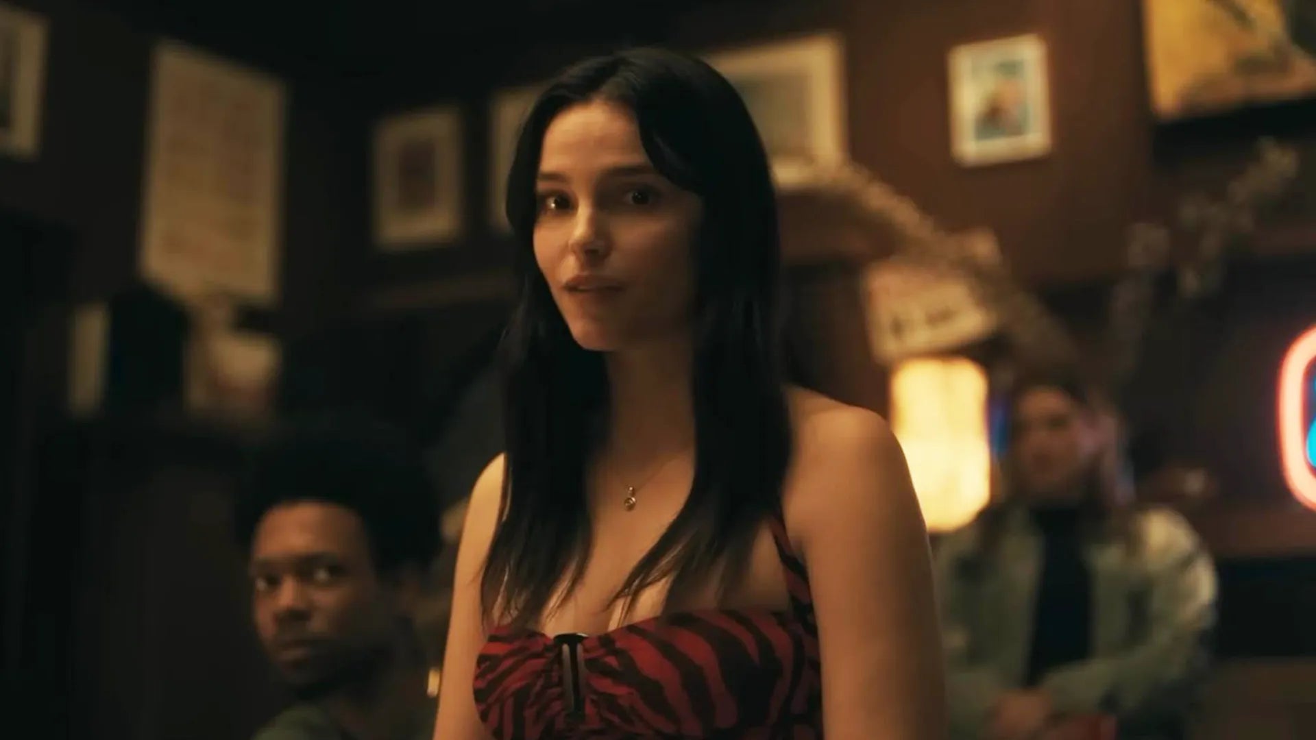 A woman with long dark hair stands in a dimly lit bar or restaurant setting, wearing a red and black patterned strapless top. Two blurred men are seated in the background.
