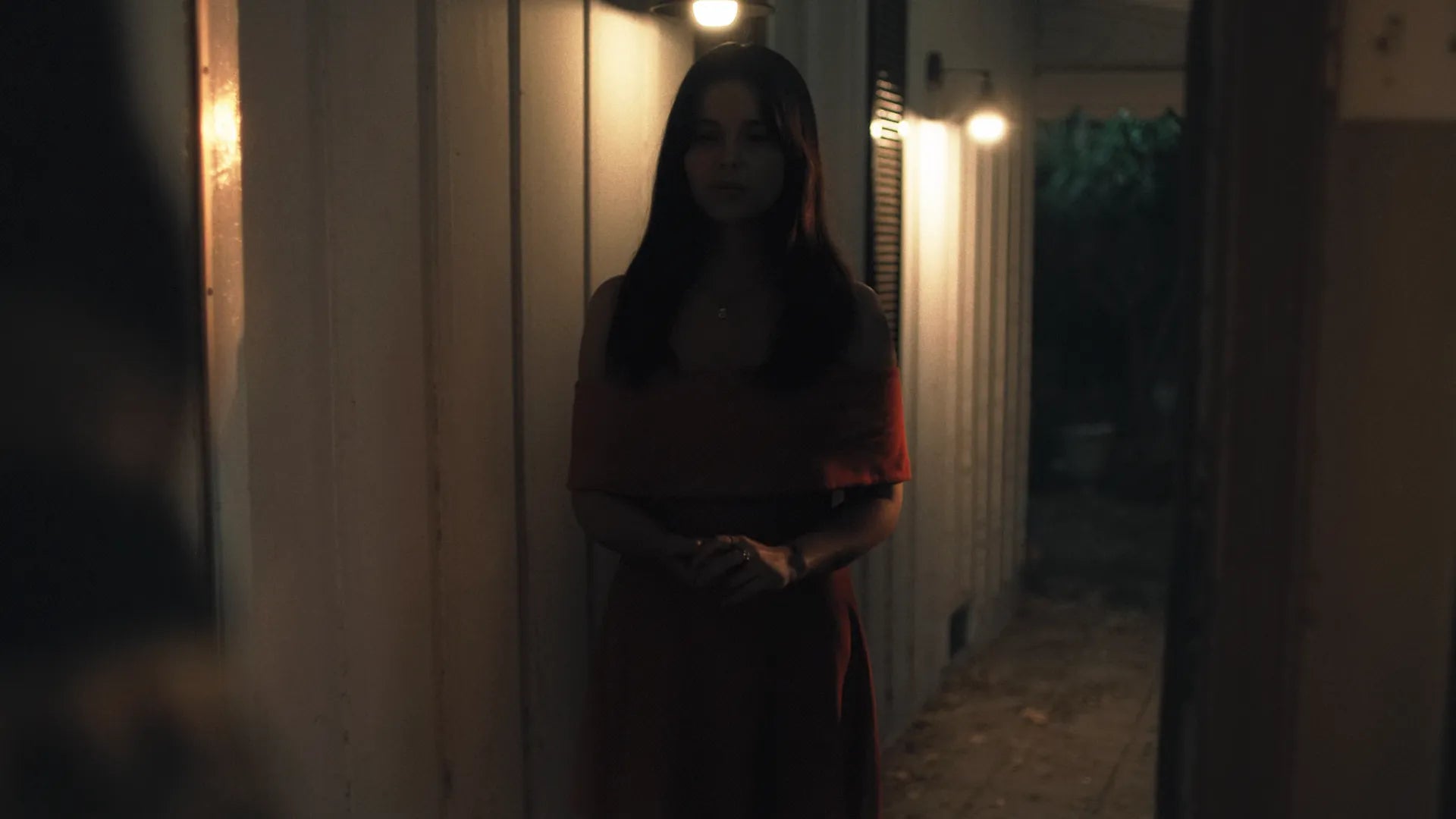 A woman with long dark hair stands in a dimly lit hallway at night, wearing a dark red dress. Warm light glows from wall-mounted bulbs on both sides of the corridor.