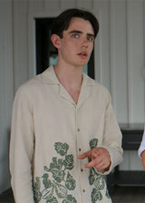 A split-screen image showing a man wearing a cream-colored button-up shirt with green floral embroidery on the left, and a studio shot of the same shirt styled with olive green pinstriped shorts on the right.
