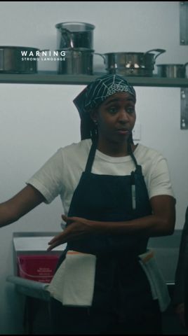 A person wearing a black apron over a white t-shirt and a patterned headscarf stands in a kitchen environment with metal pots on a shelf behind them. The text 'WARNING STRONG LANGUAGE' appears in the upper left corner.