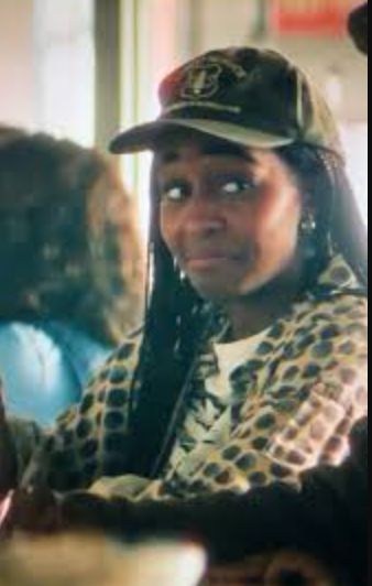 A person wearing a dark patterned cap and a leopard-print jacket, shown in a real indoor setting with another person blurred in the foreground.