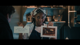 A woman in a kitchen apron and patterned headscarf holds up two printed cards or photos in a dimly lit kitchen setting. One card has text that reads, 'ANYONE CAN COOK, BUT ONLY THE FEARLESS CAN BE GREAT.