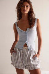 A woman modeling a light blue ribbed camisole top with a button-front overlay and striped shorts against a plain light beige studio background.